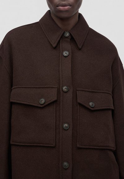 Veste en laine brun foncé avec grandes poches à rabat et boutons noirs portée par une personne visible des épaules aux lèvres.