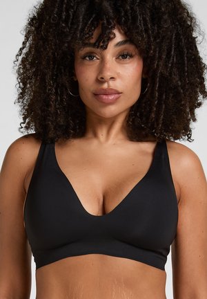 Frau mit lockigem Haar, die einen schwarzen, kabellosen BH mit V-Ausschnitt trägt, blickt frontal vor einem einfarbigen grauen Hintergrund.
