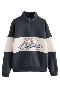 Mikina s límcem na zip, barevné bloky v námořnické modři a krémové barvě, nápis "SELF Originals 2023" v bordové a modré kurzívě. Měkká textura látky.