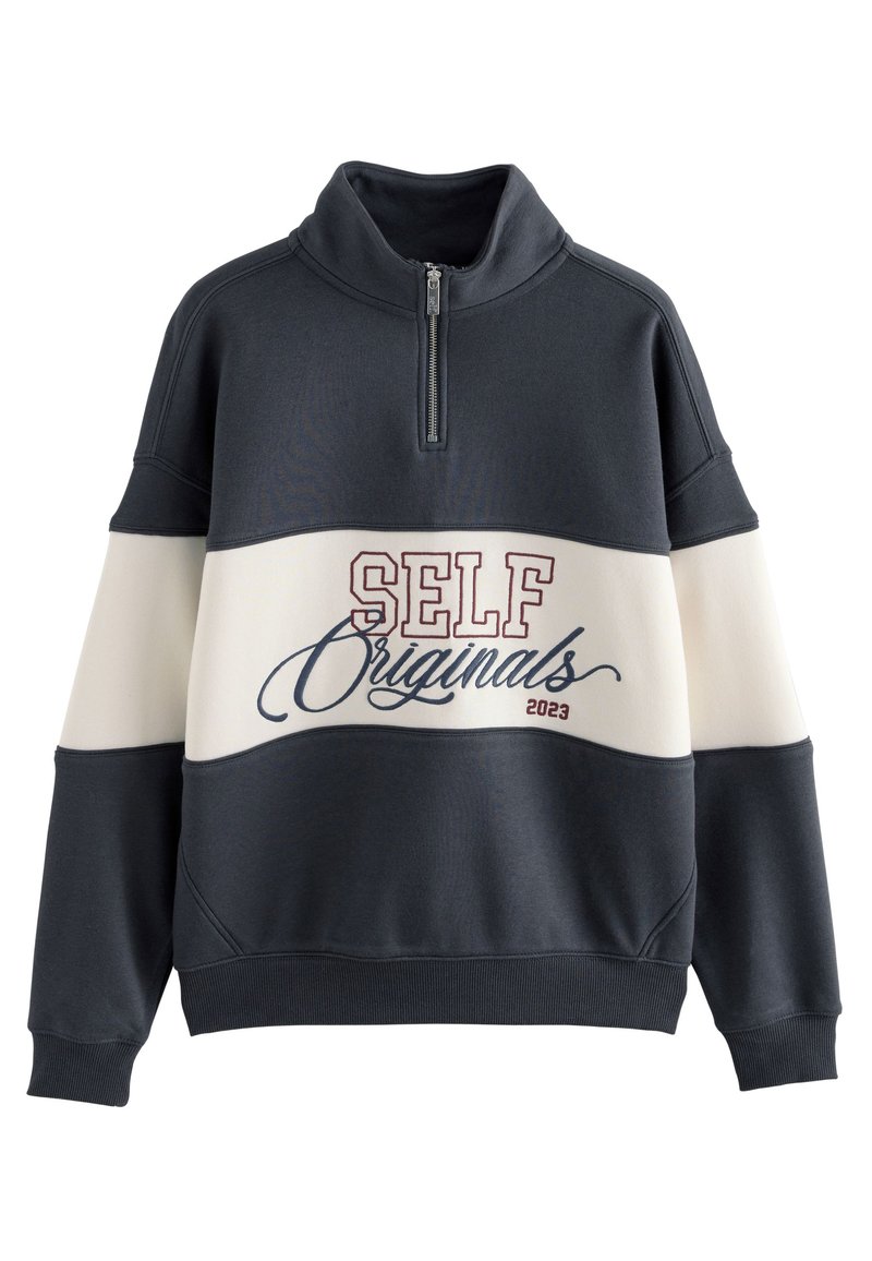Mikina s límcem na zip, barevné bloky v námořnické modři a krémové barvě, nápis "SELF Originals 2023" v bordové a modré kurzívě. Měkká textura látky.