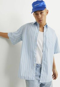 Camisa a rayas de manga corta en azul claro con botones en el frente, combinada con una camiseta blanca sin mangas y jeans azul claro. Se lleva una gorra azul.