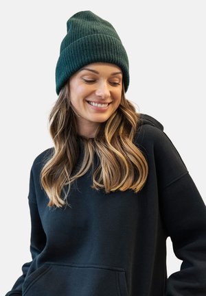 Ciemnozielona, prążkowana czapka noszona z czarnym hoodie. Hoodie ma kieszeń kangurza i luźny krój. Miękka tkanina o gładkiej fakturze.