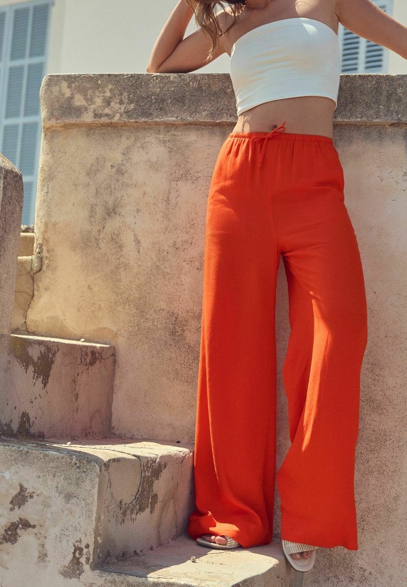 Femme appuyée contre un mur en pierre texturé, portant un top tube blanc, un pantalon large orange vif et des sandales blanches sur des marches en pierre.
