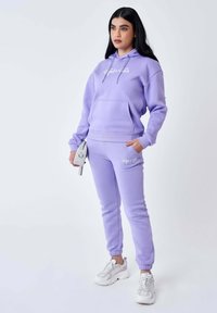 Tuta color lavanda con cappuccio, dotata di una tasca frontale e logo bianco. Include pantaloni jogger aderenti con polsini, abbinati a sneakers argentate.
