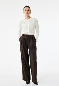 Weißer Strickcardigan mit Rundhalsausschnitt und Knopfverschluss, kombiniert mit hochgeschnittenen braunen Plisseehosen und spitzen schwarzen Absätzen.