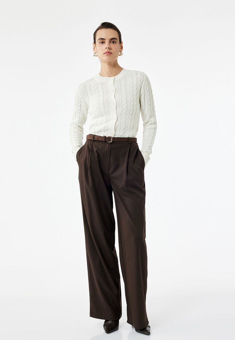Weißer Strickcardigan mit Rundhalsausschnitt und Knopfverschluss, kombiniert mit hochgeschnittenen braunen Plisseehosen und spitzen schwarzen Absätzen.