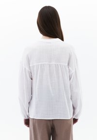 Camicia bianca leggera a maniche lunghe con tessuto testurizzato, dettaglio arricciato sul retro e scollatura rotonda. Si abbina a pantaloni beige.