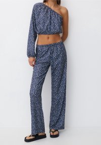 Zwei-teiliges Outfit mit einem schulterfreien, kurzen Oberteil und lockeren Hosen. Der Stoff ist blau mit kleinen Blumenmustern; beinhaltet flache Sandalen.