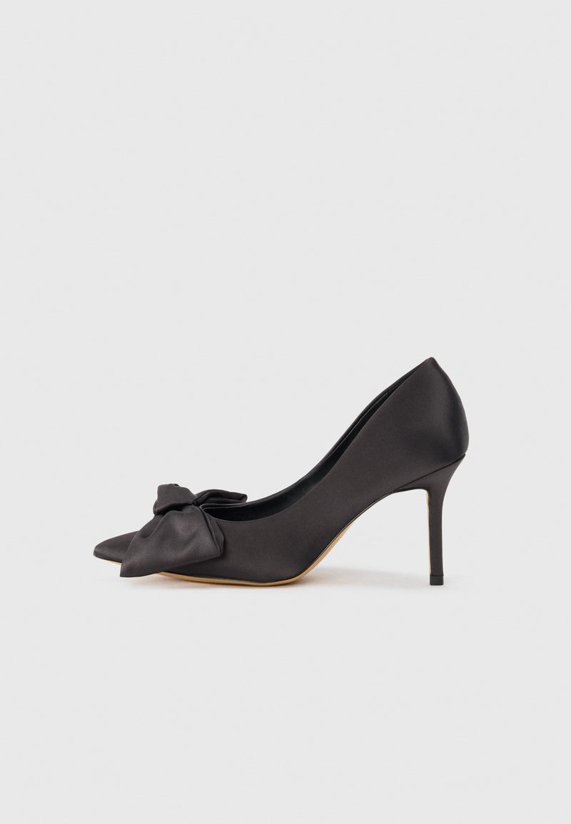 Chaussure à talon haut noire à bout pointu avec un talon stiletto et un grand nœud décoratif à l'avant, sur un fond blanc uni.