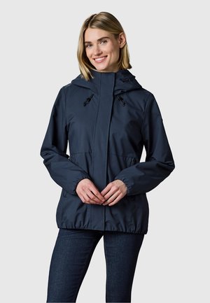 Lächelnde Frau trägt eine marineblaue Kapuzenjacke mit elastischen Bündchen und Reißverschlusstaschen, steht vor einem schlichten hellen Hintergrund.