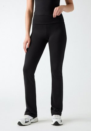 Persona che indossa pantaloni neri aderenti, top nero e scarpe da ginnastica bianche, in piedi davanti a uno sfondo chiaro e uniforme.