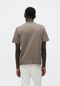 Καφέ βαμβακερό μπλουζάκι με κοντά μανίκια, στρογγυλή λαιμόκοψη και ίσιο τελείωμα. Απλό σχέδιο με άνετη εφαρμογή. Φορεμένο με ανοιχτόχρωμο παντελόνι.