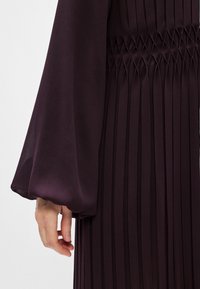 Fialová plisovaná šaty s dlhými, voľne tečúcimi rukávmi a smockovaným pásom. Tkanina má hladkú štruktúru, čím zvýrazňuje eleganciu odevu.