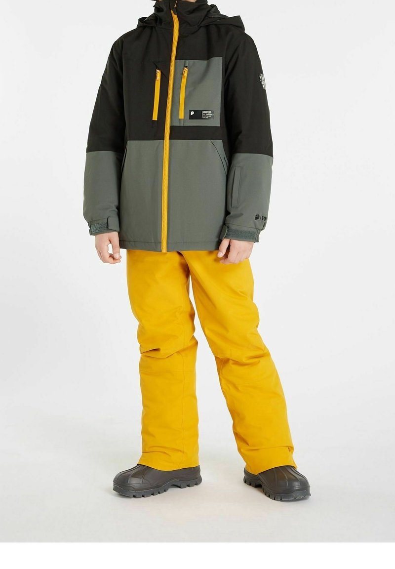 Personne portant une veste noire et grise avec une fermeture éclair jaune et des poches, un pantalon jaune vif et des bottes de neige noires, debout sur un fond blanc.