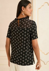 Blouse noire à manches courtes avec des motifs de nœuds blancs, présentant un panneau arrière transparent, un détail en forme de trou de serrure et un col festonné. Texture lisse.