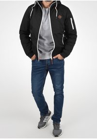 Veste noire à capuche avec fermeture éclair blanche, deux poches latérales et écusson logo. Portée sur un sweat-shirt gris, associée à un jean bleu et des baskets grises.