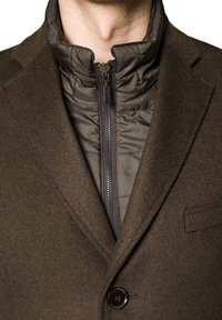 Brązowy wełniany płaszcz z wysokim kołnierzem, nałożony na ciemną pikowaną kurtkę z zamkiem. Posiada zapięcie na guziki oraz subtelną, teksturowaną tkaninę.