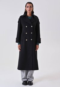 Cappotto lungo nero con design doppiopetto, grandi bottoni dorati e tasche laterali. Il modello indossa un top verde e pantaloni grigi, abbinati a stivali neri spessi.