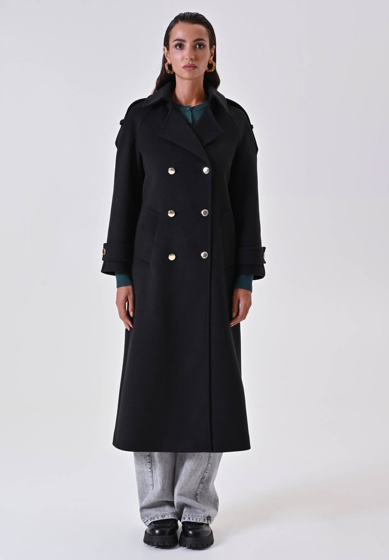 Cappotto lungo nero con design doppiopetto, grandi bottoni dorati e tasche laterali. Il modello indossa un top verde e pantaloni grigi, abbinati a stivali neri spessi.