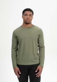 Langärmliges Hemd aus olivgrünem Baumwollstoff, mit Rundhalsausschnitt, figurschnitt, kombiniert mit schwarzen Hosen, getragen von einer Person mit kurzen Haaren.