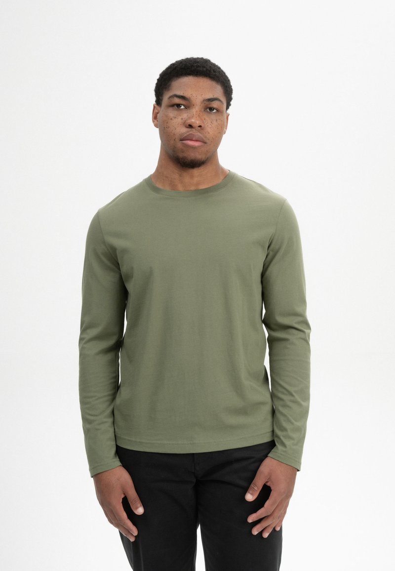 Langärmliges Hemd aus olivgrünem Baumwollstoff, mit Rundhalsausschnitt, figurschnitt, kombiniert mit schwarzen Hosen, getragen von einer Person mit kurzen Haaren.