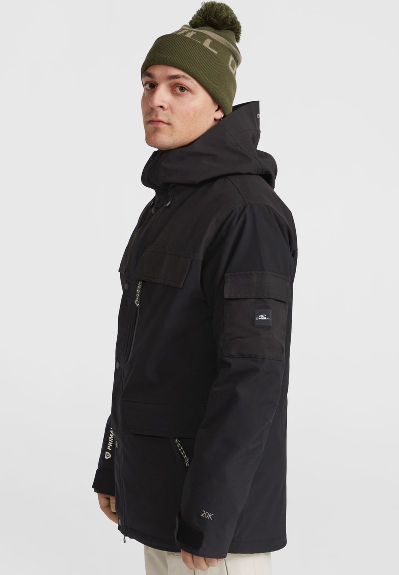 Czarna wodoodporna kurtka z kapturem, z wieloma kieszeniami, teksturowanym wykończeniem i naszywką z logo na rękawie. Noszona z oliwkową dzianinową czapką.