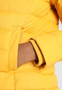 Żółta kurtka puchowa z pikowanym wzorem i ukrytą kieszenią na dłoń. Rękaw ma miękką, prążkowaną podszewkę w pasującym odcieniu żółtego.