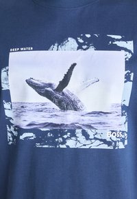 Tumši zila kokvilnas T-krekls ar vaļa grafiku taisnstūra printā, uz kura ir uzraksts "DEEP WATER" un strukturēts, abstrakts dizains baltā krāsā.