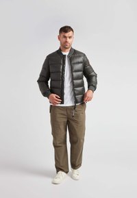 Giacca bomber imbottita verde scuro con chiusura a zip, polsini a coste e un accento rosso sulla manica, abbinata a pantaloni beige e sneakers bianche.