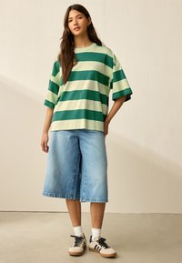 Tricou supradimensionat cu dungi verzi și crem, asortat cu șort albastru din denim cu picioare largi și adidași. Ținuta prezintă elemente de design casual și relaxate.