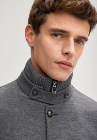 Szary sweter z dzianiny z wysokim kołnierzem, wyposażony w zamek błyskawiczny i dwa ciemne guziki. Faktura jest gładka z lekko prążkowanym wzorem.