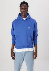Blaues Kapuzensweatshirt mit einer vorderen Tasche, mit einem Logo auf der Brust. Getragen über einem weißen Hemd, kombiniert mit hellblauen Jeans.