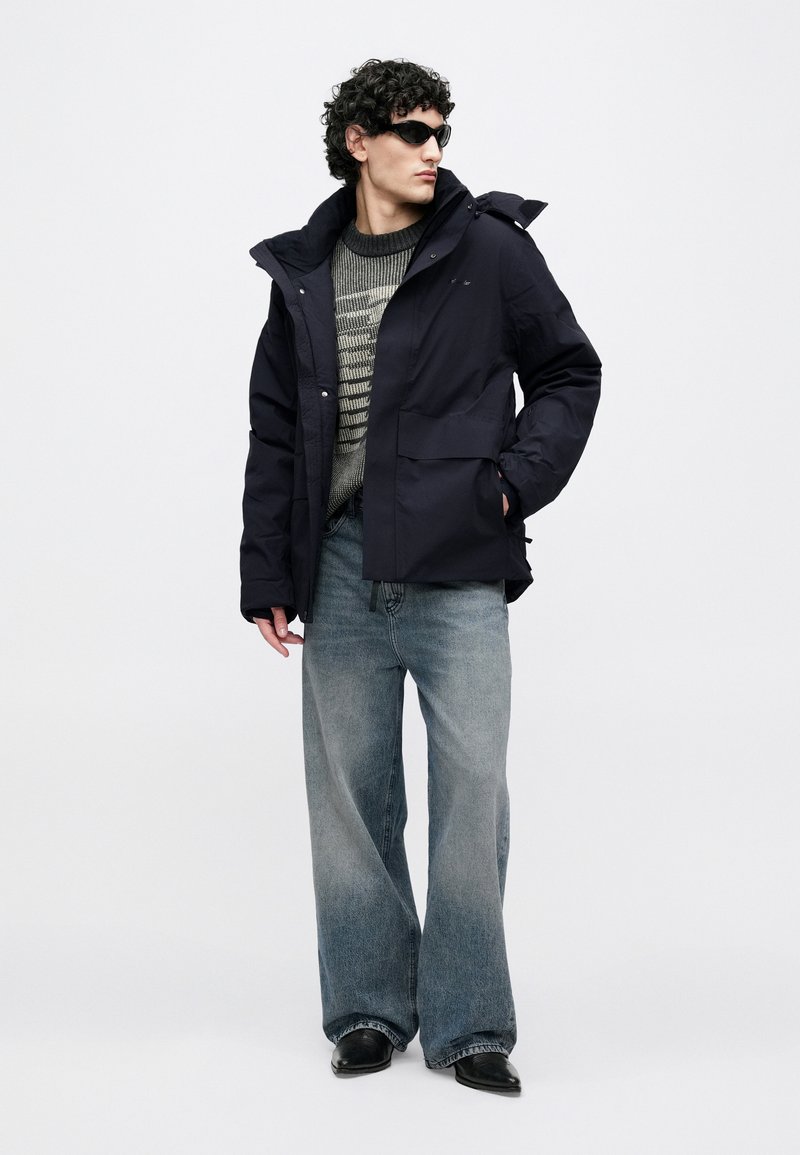 Navy insulated jacket with a hood, two front pockets, and a zipper; grey striped sweater underneath; wide-leg faded jeans; black shoes.