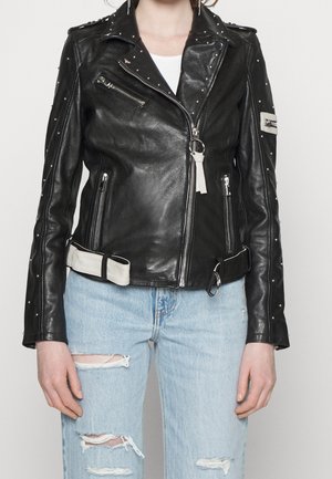 Nainen pukeutuneena mustaan niitattuun nahkaiseen biker-takkiin, jossa hopeanväriset vetoketjut ja valkoinen vyöyksityiskohta, yhdistettynä vaaleansinisiin revittyihin farkkuihin.