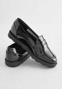 Svarta lackläderslitter med en slät yta, dekorativ båge och en texturerad sula. Har rund tå och är av typ slip-on.