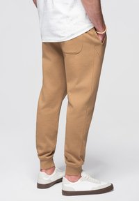 Khaki jogger kalhoty vyrobené z měkké tkaniny s bočními kapsami a zúženými manžetami, v kombinaci s bílými teniskami s hnědými podrážkami.