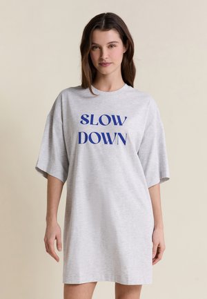 Młoda kobieta w luźnej, jasnoszarej sukience z napisem „SLOW DOWN” w kolorze niebieskim, stojąca na tle jednolitego beżowego tła.