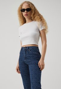 Bílý proužkovaný crop top v kombinaci s tmavě modrými džíny se geometrickým vzorem. Modelka má na sobě černé úhlové sluneční brýle. Celkově hladká textura.