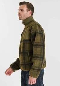 Olijfgroen fleecejack met zwart ruitpatroon, hoge kraag en een ritszak aan de voorkant. Heeft een gestructureerde stof en aansluitende mouwen.