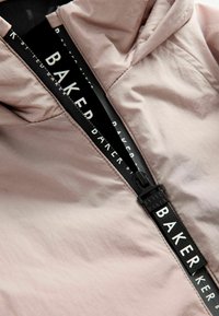 Ljusrosa jacka med dragkedja framtill och en framträdande svart dragkedja märkt "Baker", som har en slät textur och huva.