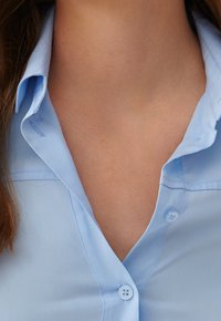 Camisa azul clara con cuello puntiagudo, que presenta una botonera y una textura suave y tejida. La tela parece ligera y transpirable.