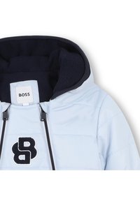 Doudoune bleu clair avec capuche marine, ornée d'un logo brodé noir et d'une fermeture éclair. Le tissu est lisse avec un design matelassé.