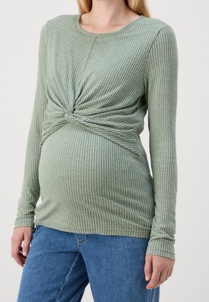 Femme enceinte portant un haut côtelé vert clair à manches longues avec un détail noué à l'avant et un jean bleu, montrée des épaules aux hanches.