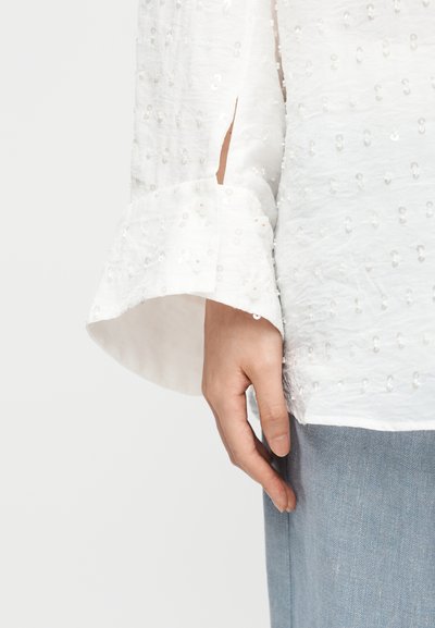 Mână relaxată pe lateral, purtând o bluză albă cu mâneci lungi texturată cu paiete, cu manșete evazate și pantaloni bleu deschis.