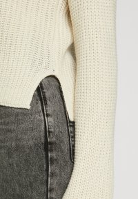 Maglione a maglia di colore crema con motivo testurizzato, caratterizzato da un taglio laterale sottile, abbinato a jeans in denim neri a vita alta.
