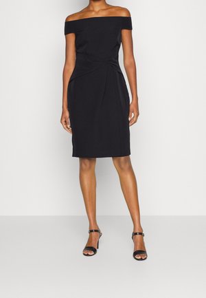 Femme portant une robe noire mi-longue épaules dénudées avec un détail plissé à la taille et des sandales noires à talons avec bride à la cheville.