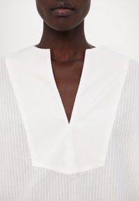 Blouse en coton blanc avec un col en V et une texture à rayures verticales, dotée d'un col audacieux et de lignes épurées.
