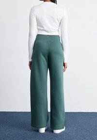 Zelené široké tepláky s jemnou texturou, elastickým pasem a bočními rozparky, spojené s přiléhavým bílým tričkem s dlouhým rukávem.