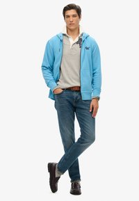 Sudadera con cremallera de color azul claro sobre una camiseta polo gris, jeans azules y zapatos marrón oscuro. Presenta bolsillos y un ajuste casual.
