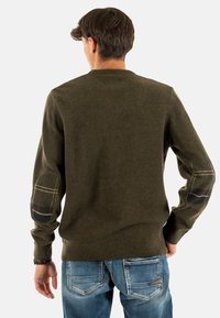 Groene gebreide trui met geribbelde onderkant en mouwen. Heeft stoffen elleboogpatches in een ruitpatroon. Draagt met blauwe jeans. Achteraanzicht.